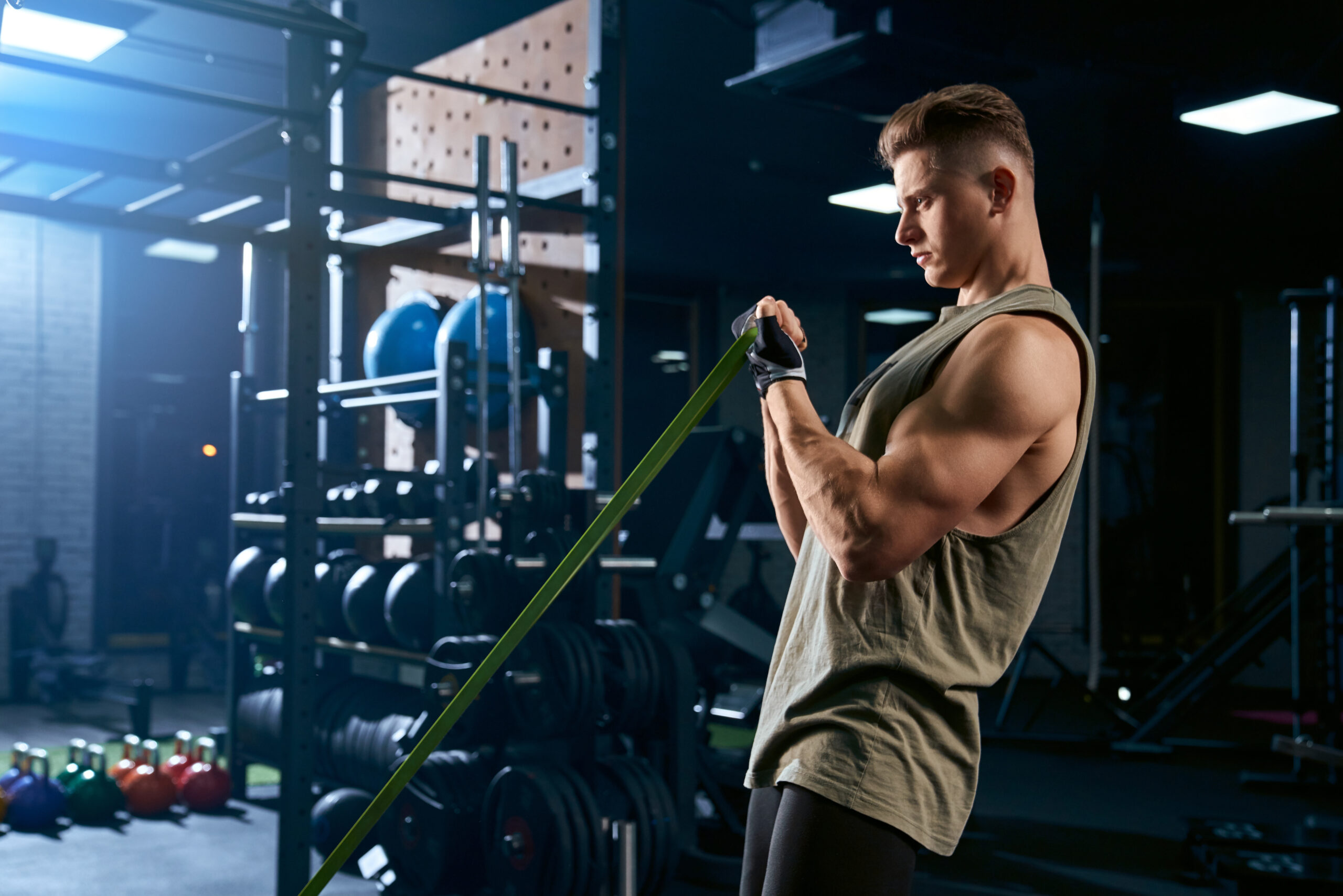 Portrait of muscular bodybuilder training arms with fitness resistance bands. Side view of man with perfect body training biceps in gym in dark atmosphere. Concept of bodybuilding.